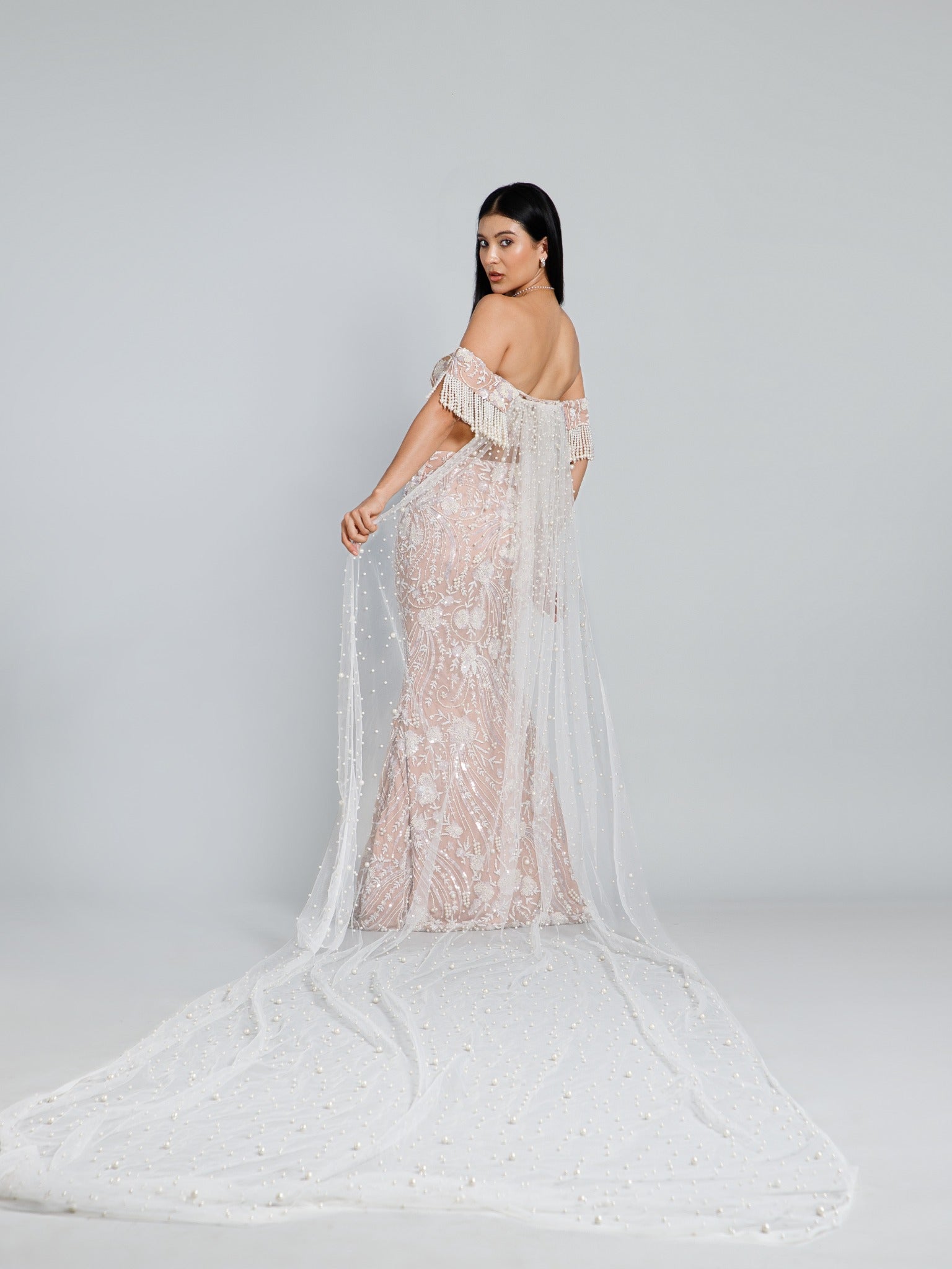 Back view of the beige embroidered fish cut lehenga by DEEPANSHI MASSEY LABEL, featuring a fitted silhouette and a long sheer cape adorned with scattered pearls.