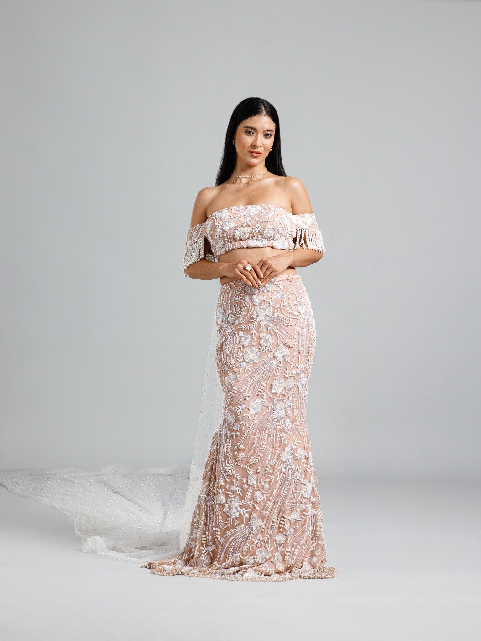 Full-length view of the beige fish cut lehenga by DEEPANSHI MASSEY LABEL, featuring intricate floral embroidery, an off-shoulder pearl-fringed blouse, and a trailing pearl-embellished cape.
